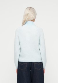 Gaiši zils, mīksts adījums džemperis ar augstu apkakli un ševrona rakstu, apvienots ar tumšiem džinsu platgurniem, redzams aizmugures skats.