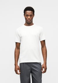 Λευκό βαμβακερό t-shirt με κοντά μανίκια και στρογγυλή λαιμόκοψη. Διαθέτει διακριτική σήμανση στο στήθος. Συνδυάζεται με γκρι παντελόνια.