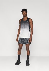 Αθλητικό αμάνικο μπλουζάκι σε κλίση μαύρου προς λευκού με αφηρημένο σχέδιο; συνδυασμένο με μαύρες βερμούδες με σχέδιο και μαύρα υποδήματα, συμπληρωμένο από σκούρες κάλτσες.