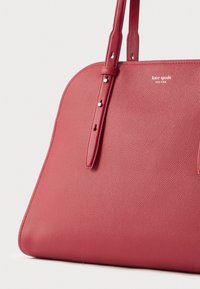 Sarkana ādas soma ar teksturētu virsmu, izliekta forma, divas pleca siksnas un reljefā iegravēts logotips priekšpusē. Sudraba krāsas metāla elementi.