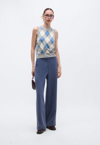 Μπλε και κρεμ τοπ χωρίς μανίκια με σχέδιο αργκλάι και γκρι ριμπ τελείωμα, σε συνδυασμό με φαρδιά, ναυτικά παντελόνια και σκούρα παπούτσια, κρατώντας μια μικρή τσάντα.