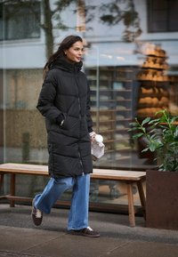 Schwarze gefütterte Jacke mit Kapuze, Reißverschluss und Nahtsteppungen; blaue weit geschnittene Jeans; braune Sneakers; hält einen Becher und eine Papiertüte.