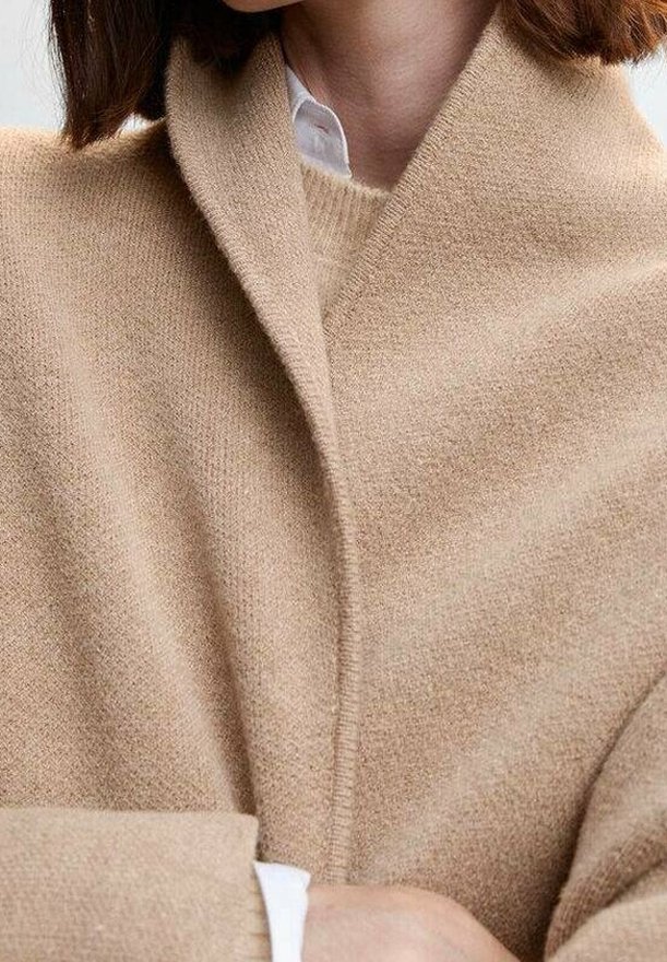 Femme portant un manteau en laine beige par-dessus un pull crème et une chemise blanche à col, les bras croisés à la taille.