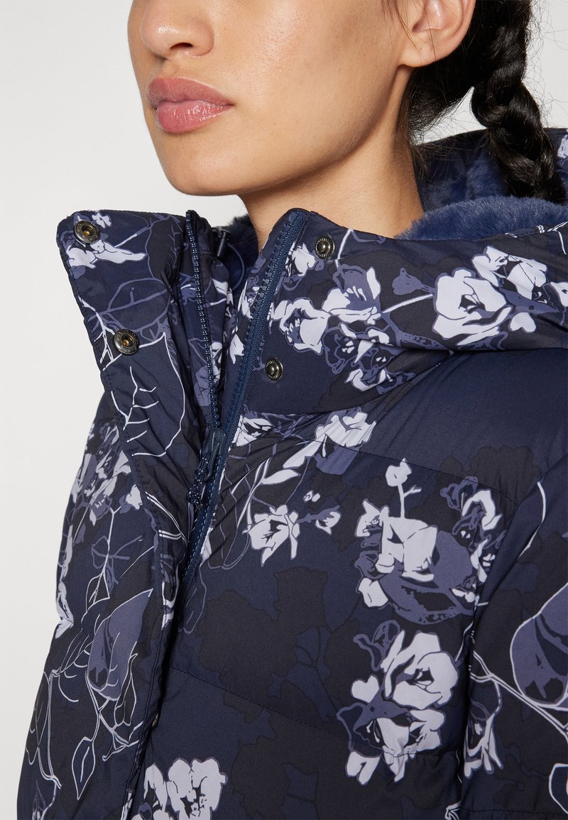 Doudoune florale bleu marine avec un col haut, fermeture éclair et boutons-pression. Présente des motifs floraux abstraits blancs et une doublure douce.
