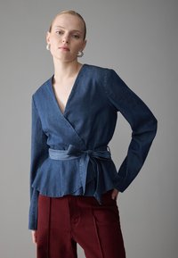 Blusa in denim avvolgente con maniche lunghe, scollatura a V profonda, orlo peplum, vita legata, in una tonalità di blu scuro, abbinata a pantaloni bordeaux.