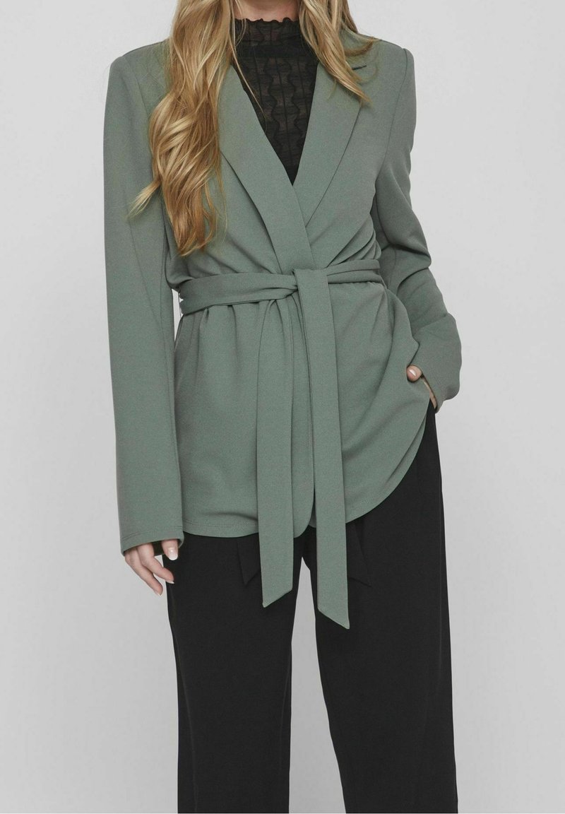 Grön kavaj med långa ärmar, ett knytbälte i midjan och slätt tyg. Lageröver en svart strukturerad blus. Svarta byxor.