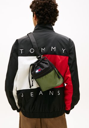 Persona que lleva una chaqueta Tommy Jeans en negro, blanco y rojo, con un bolso bandolera Tommy Jeans en negro y verde.