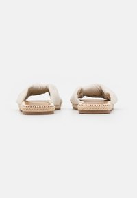 Een paar beige platte slip-on sandalen met gevoerde bandjes en geweven juten tussenzolen, gezien van voren op een witte achtergrond.