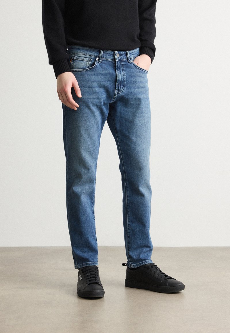 Jean bleu clair à coupe droite, en denim, avec un design à cinq poches et un léger effet de décoloration, associés à des baskets noires.