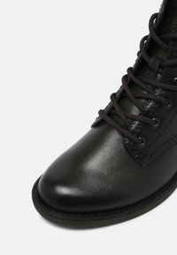 Botte noire en cuir à lacets avec bout arrondi et semelle robuste, affichant des coutures détaillées et des œillets en métal sur un fond neutre.