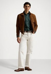 Brązowa zamszowa kurtka na ciemnozielonej koszuli, białe spodnie i brązowe skórzane buty. Model stoi z luźno opuszczonymi ramionami, prezentując strój.