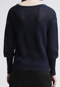 Tmavě modrý pletený svetr s texturovaným, vzdušným vzorem nahoře a jednobarevnou spodní částí. Má volný střih a široký výstřih.