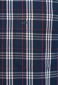 Granatowy materiał w kratę z białymi i czerwonymi liniami, z przeszywaną kieszenią na piersi i małym, haftowanym logo biegnącego konia.