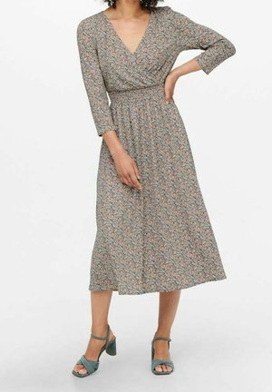 Femme portant une robe midi fleurie à manches longues et col en V, avec une taille froncée et des sandales à talons blocs ouvertes sur les orteils de couleur bleu sarcelle, debout avec une main sur la hanche.