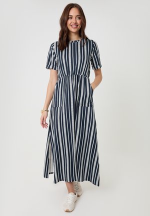 Femme portant une robe midi à rayures verticales bleu marine et blanches, manches courtes, et des baskets blanches, debout avec une main dans la poche.