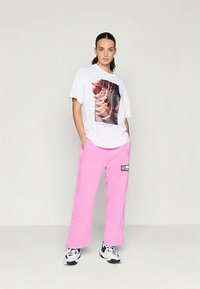 Vrouw staat met handen in de zakken, gekleed in een wit graphic t-shirt, felroze wijde broek met zijknopen en zwart-witte sneakers.