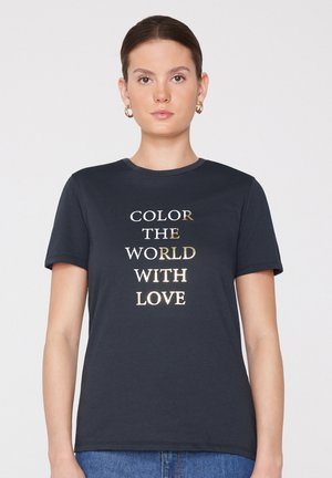Kobieta z ciemnymi, związanymi włosami nosi złote kolczyki koła i granatową koszulkę z tekstem w kolorze złotym i białym "COLOR THE WORLD WITH LOVE."