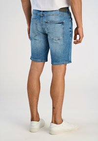 Niebieskie szorty dżinsowe o klasycznym kroju, postrzępionym końcu i pięciu kieszeniach. Widok z tyłu pokazuje szwy tylnej kieszeni oraz jasny odcień.