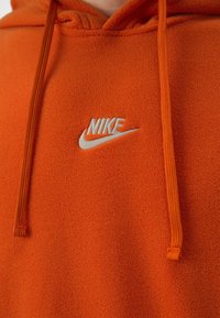 Nära bild av en orange fleecetröja med huva som visar en vit och orange broderad Nike-logotyp och matchande orange dragsnören.