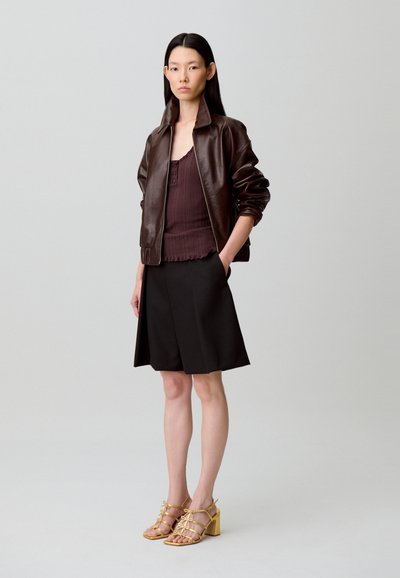 Chaqueta bomber de cuero marrón, camiseta sin mangas acanalada color granate, falda negra y sandalias de tacón amarillas con tiras, mostrando texturas y colores variados.