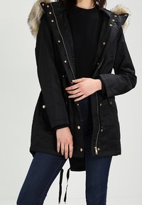 Svart parka-jacka med fuskpäls på huvan, dragkedja och tryckknappar, justerbar dragsko i midjan och två framfickor. Mjuk textur och varm design.