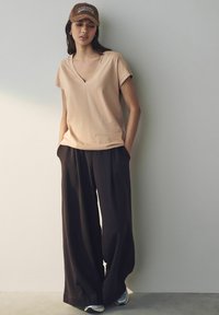 Ljus rosa t-shirt med v-ringning, avslappnad passform; löst sittande mörka byxor, mjukt material; brun keps med "AMOUR"-bokstäver; skor med grå detaljer.