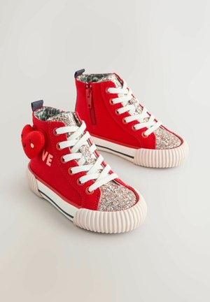 Piros magas szárú sportcipők csillogó orrésszel, fehér fűzőkkel, texturált gumitalppal, és egy mosolygós arcot ábrázoló szív alakú medállal.