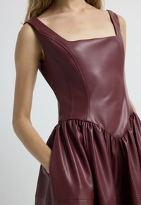 Vestido de cuero burdeos con escote cuadrado, corsé ajustado y falda acampanada con pliegues y bolsillos laterales. Textura suave y costuras sutiles.
