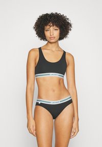 Fekete bikini szett, amely egy ujj nélküli toppal rendelkezik, csíkozott gumírozott pánttal és hozzáillő hipster alsóval, logóval a derékszíjon.