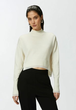 Maglione bianco corto con texture a coste e colletto alto. Abbinato a pantaloni neri con finitura liscia e cintura elastica.