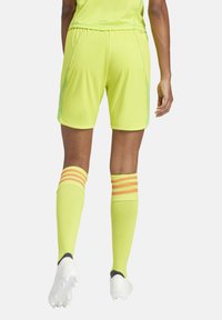 Limoeng groene sportshorts met zijpanelen van mesh, gecombineerd met geel-oranje gestreepte sokken en witte sportschoenen. Achteraanzicht.