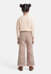 Παιδί με διπλό κότσο στα μαλλιά, φοράει κρεμ πλεκτό πουλόβερ, παντελόνι με φαρδιά μπάρα και λεοπάρ σχέδιο, και καφέ μπότες, στέκεται γυρισμένο προς τα πίσω σε λευκό φόντο.