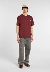 Viininpunainen lyhythihainen t-paita, jossa on rintatasku, yhdistetty harmaisiin leveälahkeisiin housuihin ja ruskeisiin puukenkäihin, neutraalia taustaa vasten.