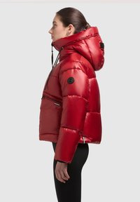 Chaqueta de plumas roja con un acabado brillante, que cuenta con capucha, bolsillo en el pecho con cremallera y parche negro con logo en la manga.