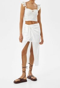 Top corto blanco de canalé con mangas acampanadas y lazo frontal, combinado con una falda envolvente blanca con una abertura alta y sandalias marrones con tiras.