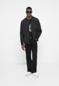 Sort bomberjakke med et let design, båret over en sort grafisk T-shirt, parret med sorte jeans og sneakers med beige detaljer.