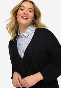 Cardigan nero in maglia con un motivo testurizzato, dotato di scollo a V profondo, chiusura con bottoni e maniche lunghe, indossato sopra una camicia a righe con colletto.