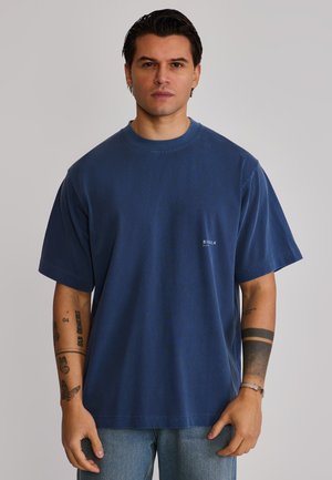 Mand med mørkt hår iført en ensfarvet, blå, oversized T-shirt og lyseblå jeans, der viser tatoveringer på begge underarme mod en grå baggrund.