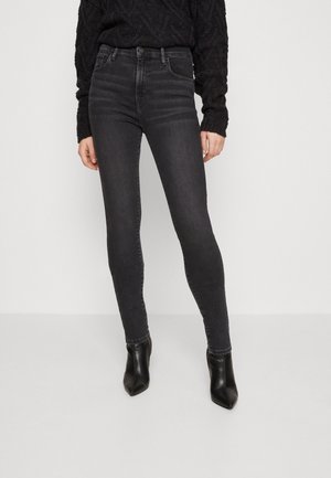 Frau trägt schwarzen Kabelstrickpullover, hoch taillierte dunkelgraue Skinny-Jeans und schwarze Knöchelstiefel, steht vor einem schlichten hellen Hintergrund.