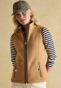 Beige Fleecewesten mit braunem Reißverschluss, zwei Fronttaschen, getragen über einem navy- und weiß gestreiften Langarmshirt, mit einer gemusterten Mütze.