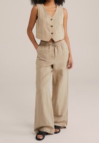 Gilet corto in lino beige con bottoni, abbinato a pantaloni larghi leggeri beige con vita elastica e tasche laterali. Sandali neri.