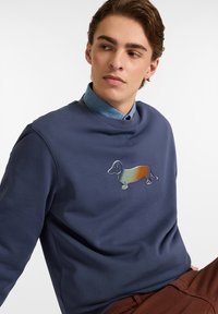 Marineblaues Sweatshirt mit rundem Halsausschnitt, das ein farbenfrohes gesticktes Design eines Dackels auf der Brust zeigt. Aus weichem Stoff gefertigt, kombiniert mit einem blauen Hemd.