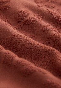Tessuto terracotta con un motivo strutturato e in rilievo. Texture morbida e tessuta con una finitura leggermente vellutata. Tono caldo e terroso con una sottile profondità.