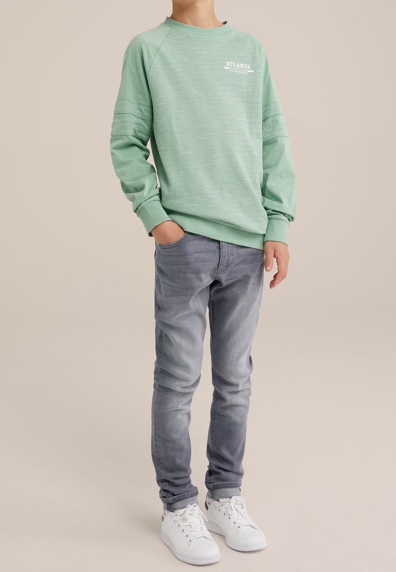 Světle zelená mikina s žebrovanými manžetami a kulatým výstřihem, s bílým textem "ATLANTA". V kombinaci s úzkými šedými džíny a bílými teniskami.