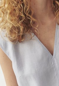 Gaiši zils lina tops ar V veida kakla izgriezumu un īsām piedurknēm. Audums izskatās mīksts, ar nelielu spīdumu un minimālām šuvju detaļām.