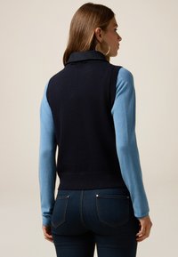 Marineblauwe en lichtblauwe mouwloze gebreide trui met een kraag en geribbelde zoom, gecombineerd met donkerdenim jeans. Eenvoudig ontwerp, soepele textuur.