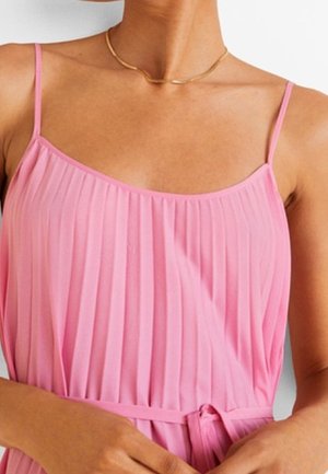 Frau trägt ein ärmelloses, rosa plissiertes Kleid mit Spaghettiträgern und eine dünne goldene Halskette, die Hände ruhen nahe der Taille.