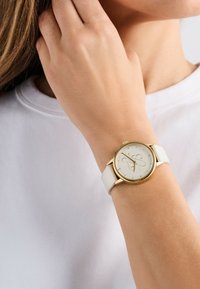 Orologio con cinturino in pelle bianca, cassa oro, quadrante rotondo e dettagli in rilievo. Il cinturino appare liscio e flessibile.