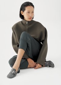 Übergroßer dunkelgrüner Pullover mit hohem Kragen und Reißverschluss, kombiniert mit dunkelblauen, schmal zulaufenden Hosen und grauen, blockabsatz Schuhen mit Schnalle.