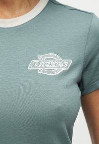 Κοντινό πλάνο ενός ατόμου που φοράει μπλούζα σε χρώμα θαλασσινό πράσινο με λευκό λογότυπο "Dickies" και σχέδιο πετάλου στο στήθος.